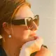 Foto de perfil da aluna Sarah, mulher usando óculos de sol elegantes e brincos de pérola, apoiando o queixo na mão em iluminação dourada e ensolarada.